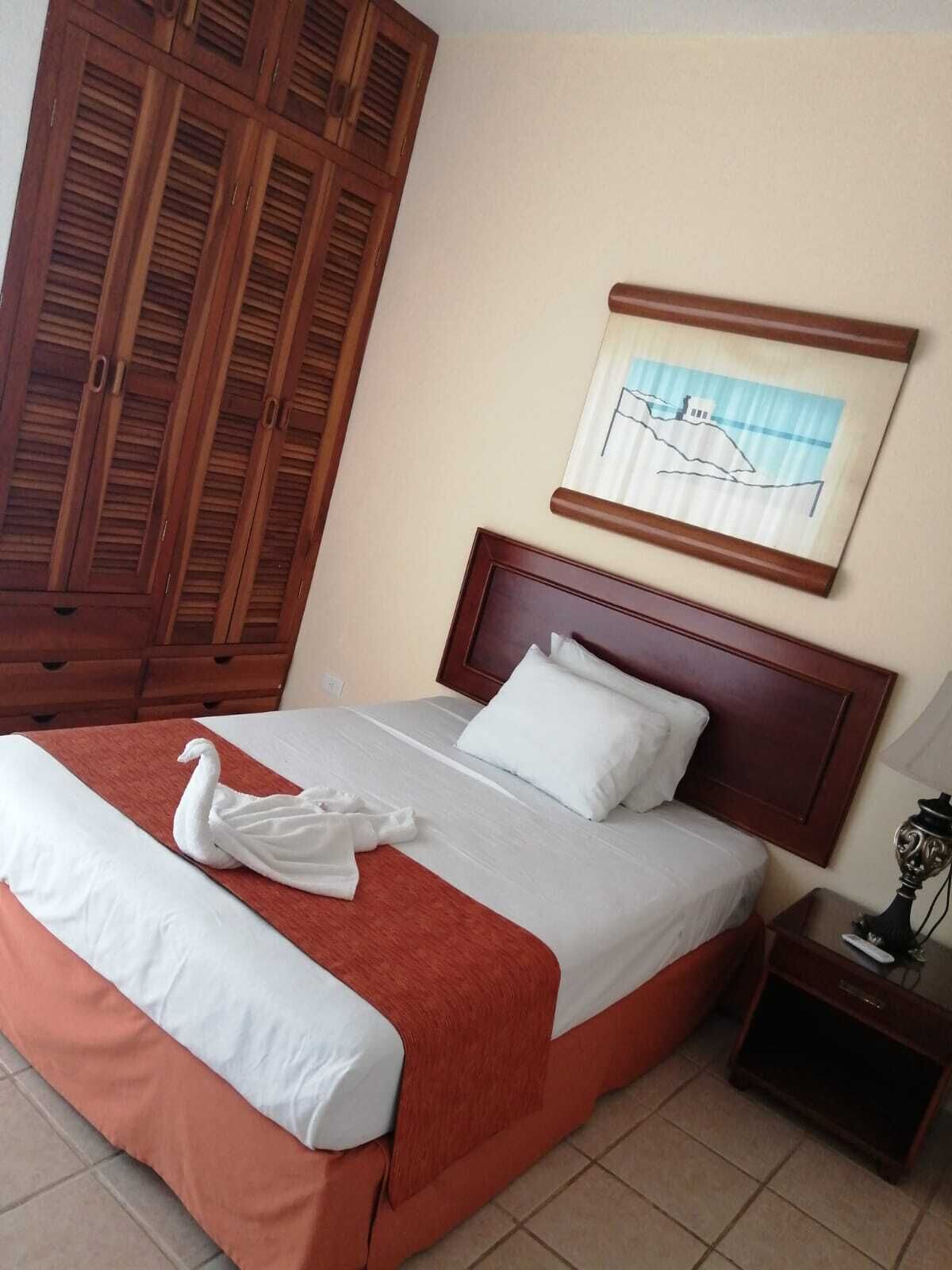A hotel room with a bed, a wooden wardrobe, a framed picture on the wall, and a nightstand with a lamp.