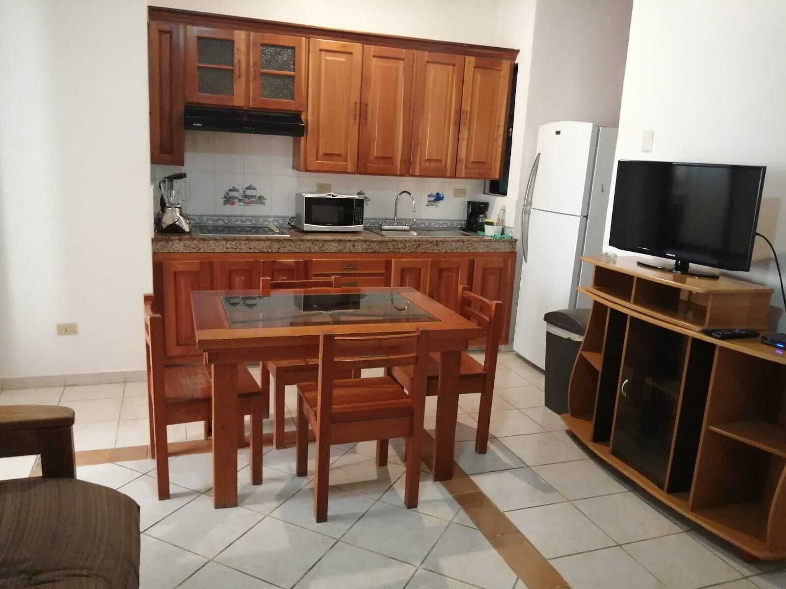 A compact kitchen with wooden cabinets, a microwave, a sink, and a television on a wooden stand.