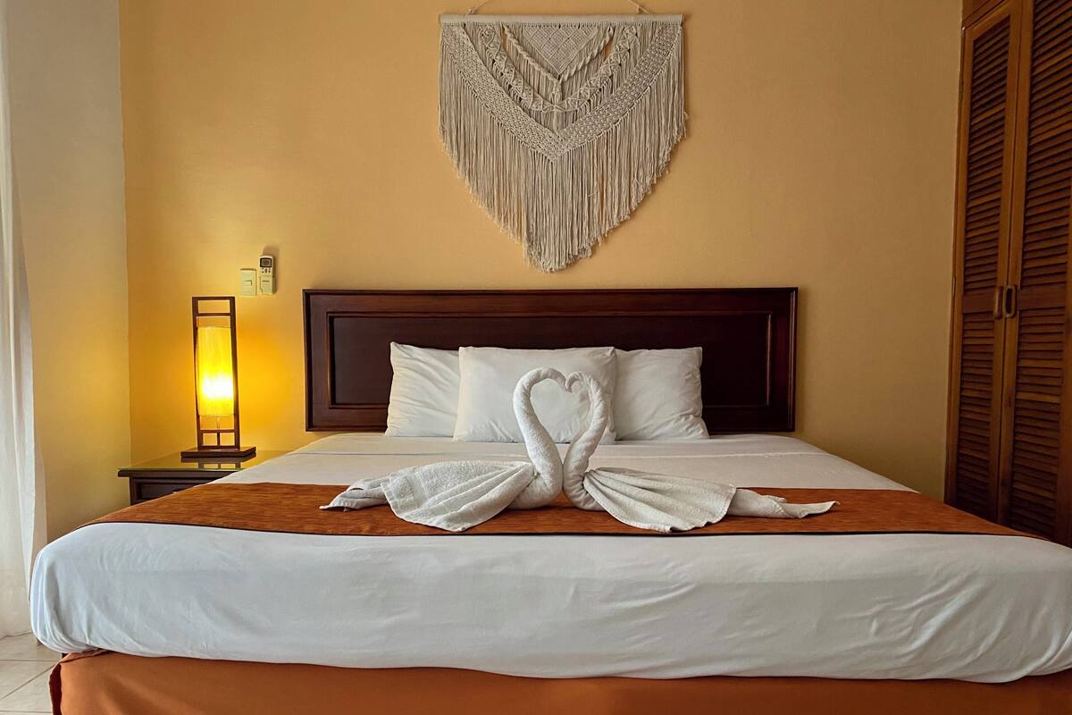 A hotel room with a bed, a wall-mounted macrame piece, a bedside lamp, and a wooden door.
