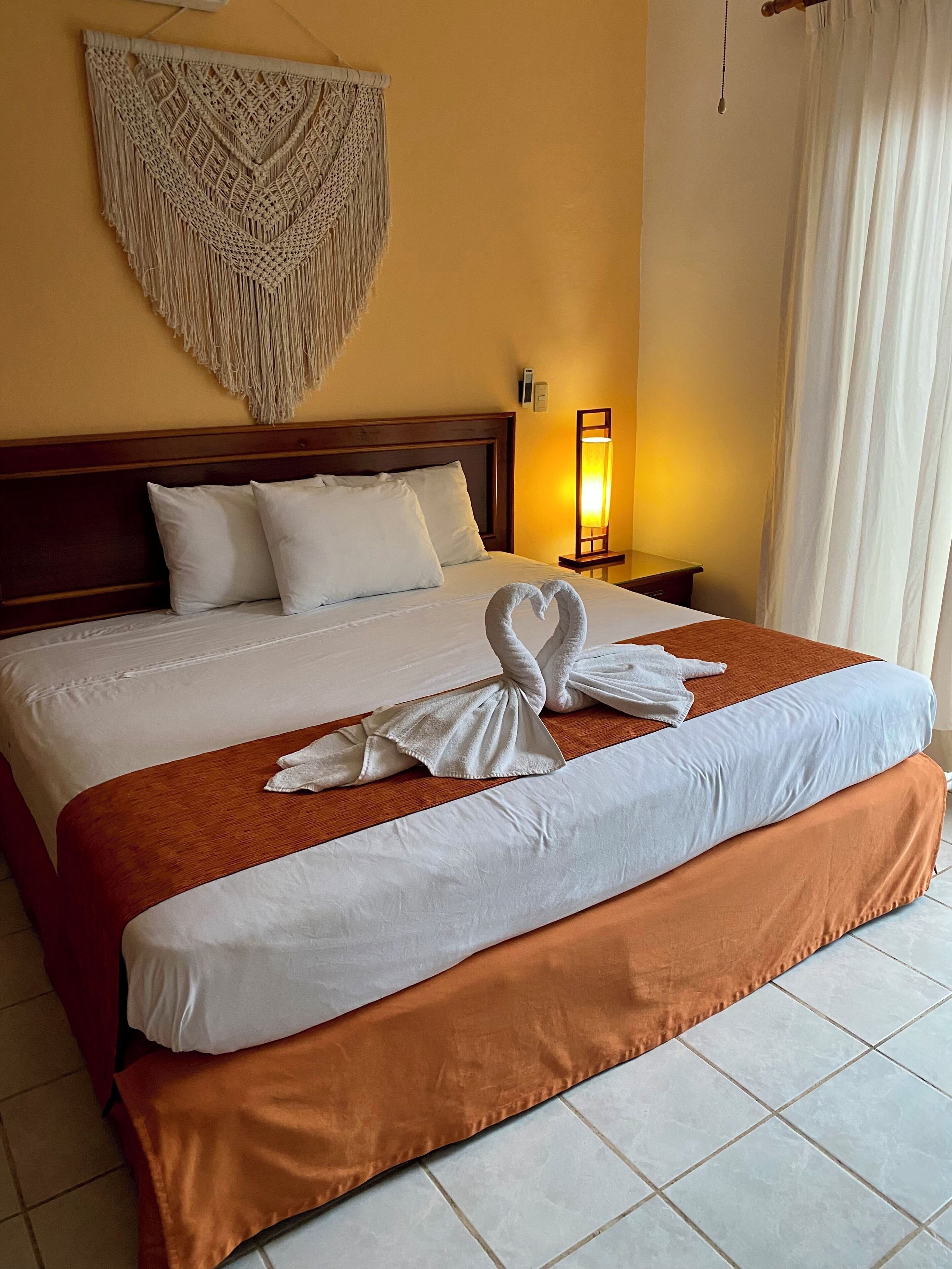 A neatly made bed with white and brown bedding, a macrame wall hanging, and a bedside lamp.