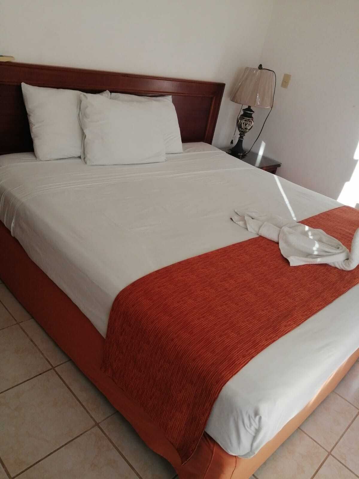 Una cama bien tendida con sábanas blancas y una manta roja, un cabecero de madera y una lámpara de noche.