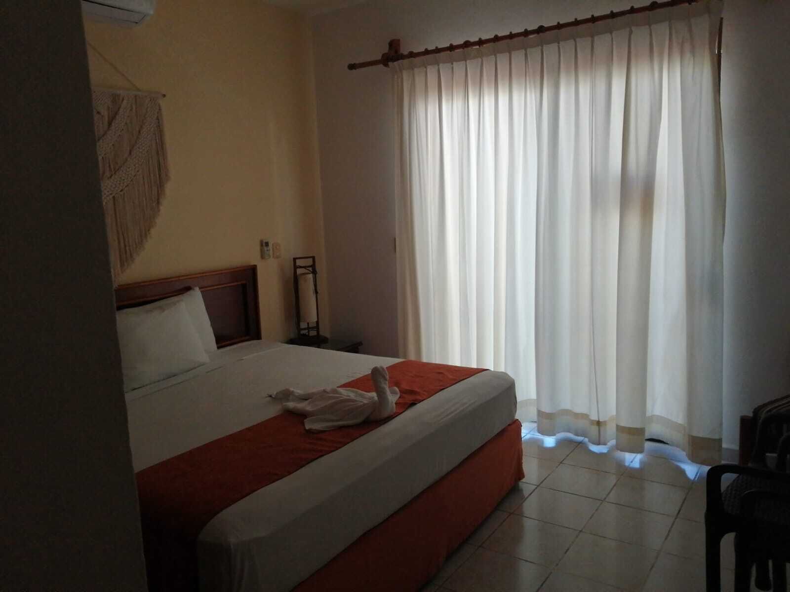 Una habitación de hotel con cama, mesita de noche, lámpara y ventana con cortinas.