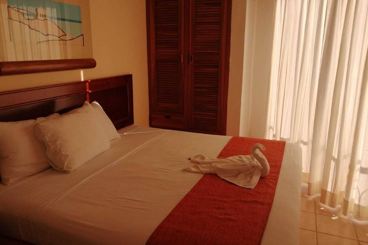 A hotel room with a bed, a wooden headboard, a red towel, and a painting on the wall.