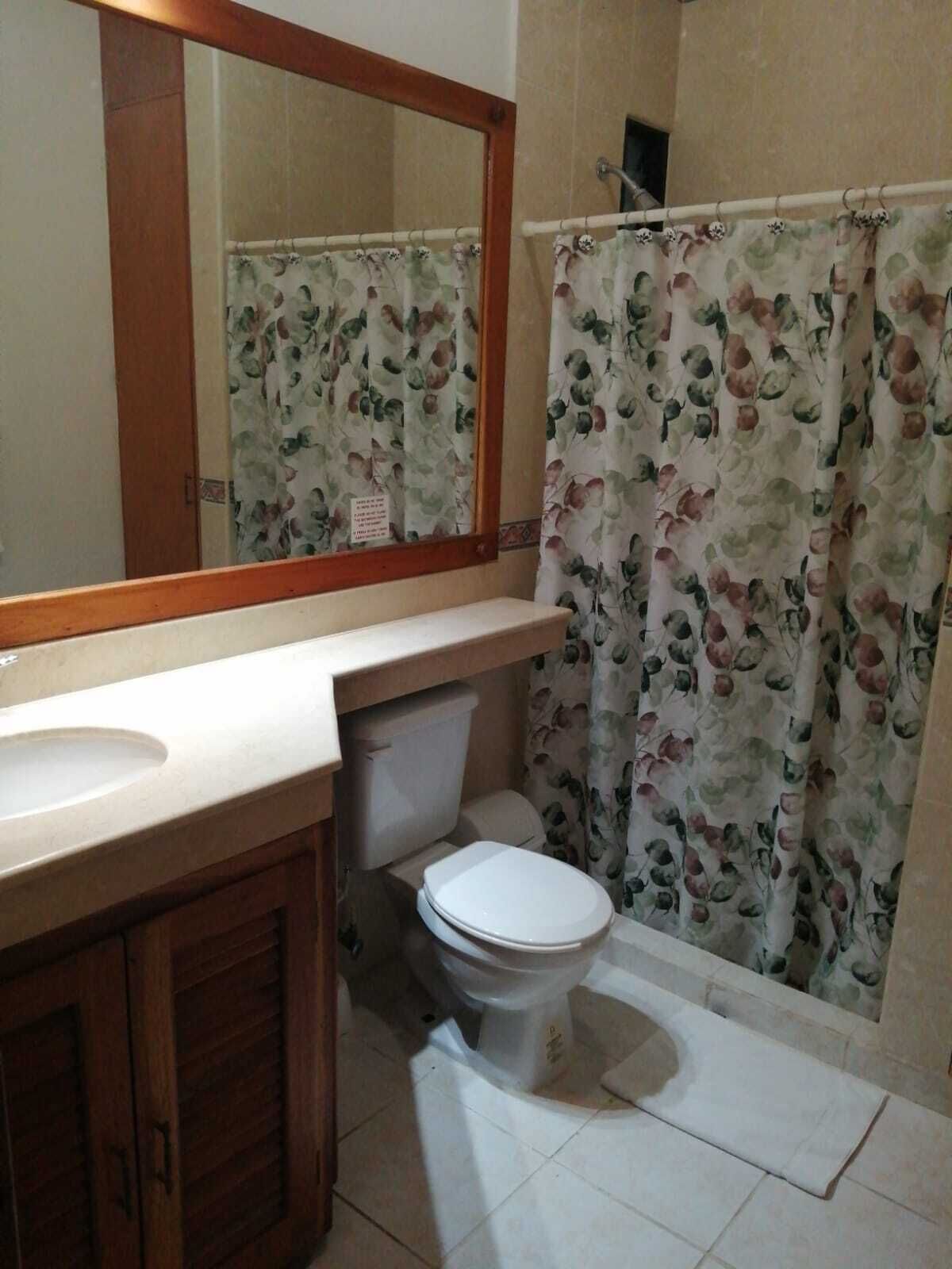 Un baño con inodoro, lavamanos, espejo y cortina de ducha estampada con motivos florales.