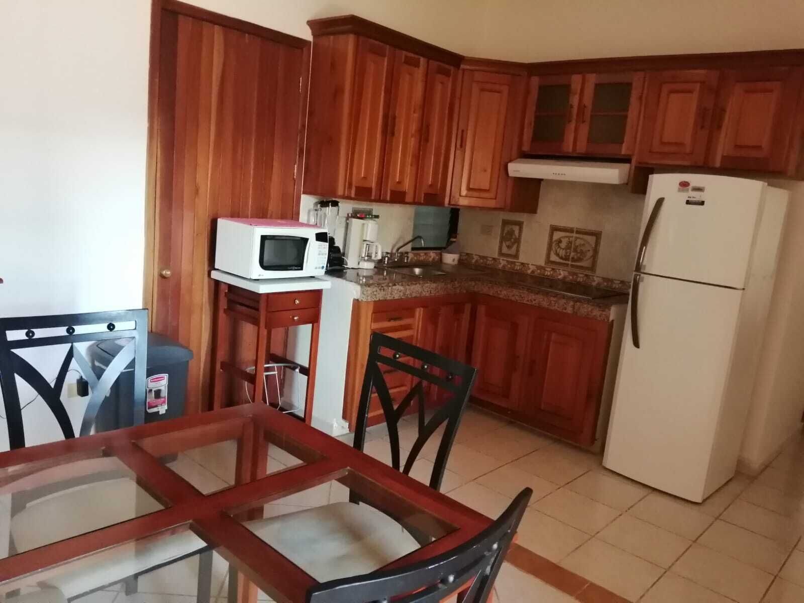 A compact kitchen with wooden cabinets, a white refrigerator, a microwave, and a dining table with chairs.