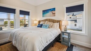 A bedroom with a large bed, two bedside tables, a nightstand, a painting on the wall, and a view through the window. - Your Fresh Beach Oasis! (Corolla)
