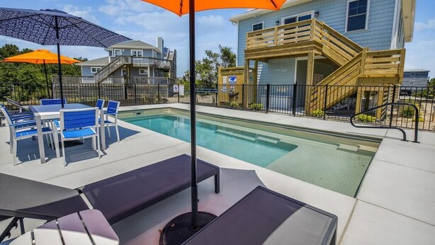 A swimming pool area with lounge chairs and umbrellas, a wooden deck with stairs, and a light blue building in the background. - Your Fresh Beach Oasis! (Corolla)