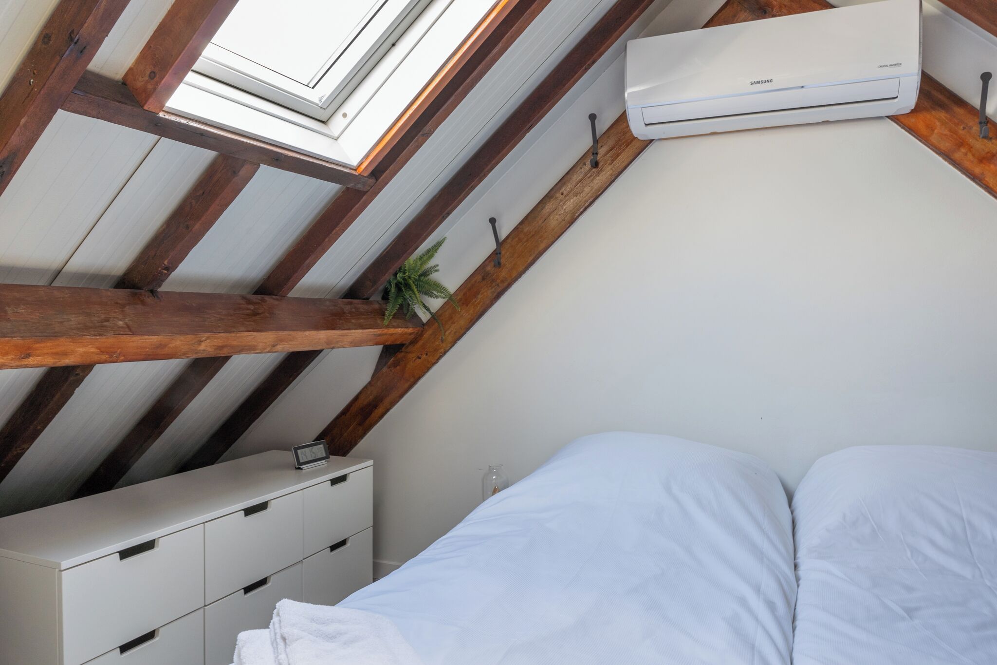 Ein Schlafzimmer mit schrägen Wänden, Holzbalken, einem weißen Bett, einem Nachttisch, einem kleinen Topfpflanzen und einer Klimaanlage.
