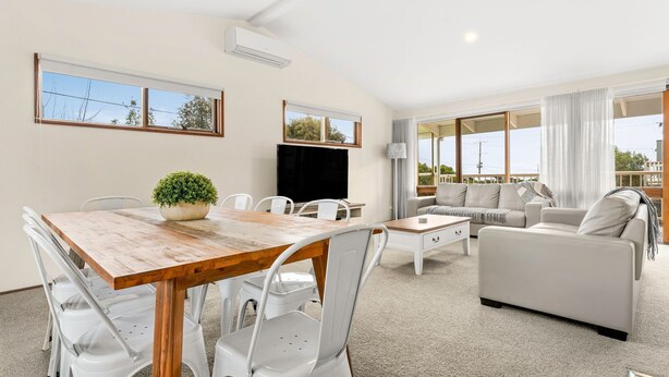 A modern living room with a wooden dining table, white chairs, a grey sofa, and a TV. - Views on Broadway - Pet friendly 4 bedroom home (San Remo)