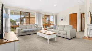 A spacious living room with two sofas, a coffee table, and a TV. Large windows offer a view of a brick building and greenery. - Views on Broadway - Pet friendly 4 bedroom home (San Remo)