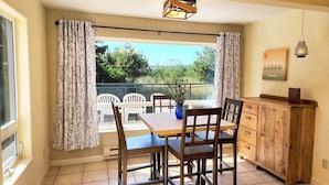 A dining area with a wooden table and chairs, a blue vase with flowers, a wooden cabinet, and a view of greenery outside. - Fisherman's Delight in Waldport! Dog Friendly! Hot tub! (Waldport)