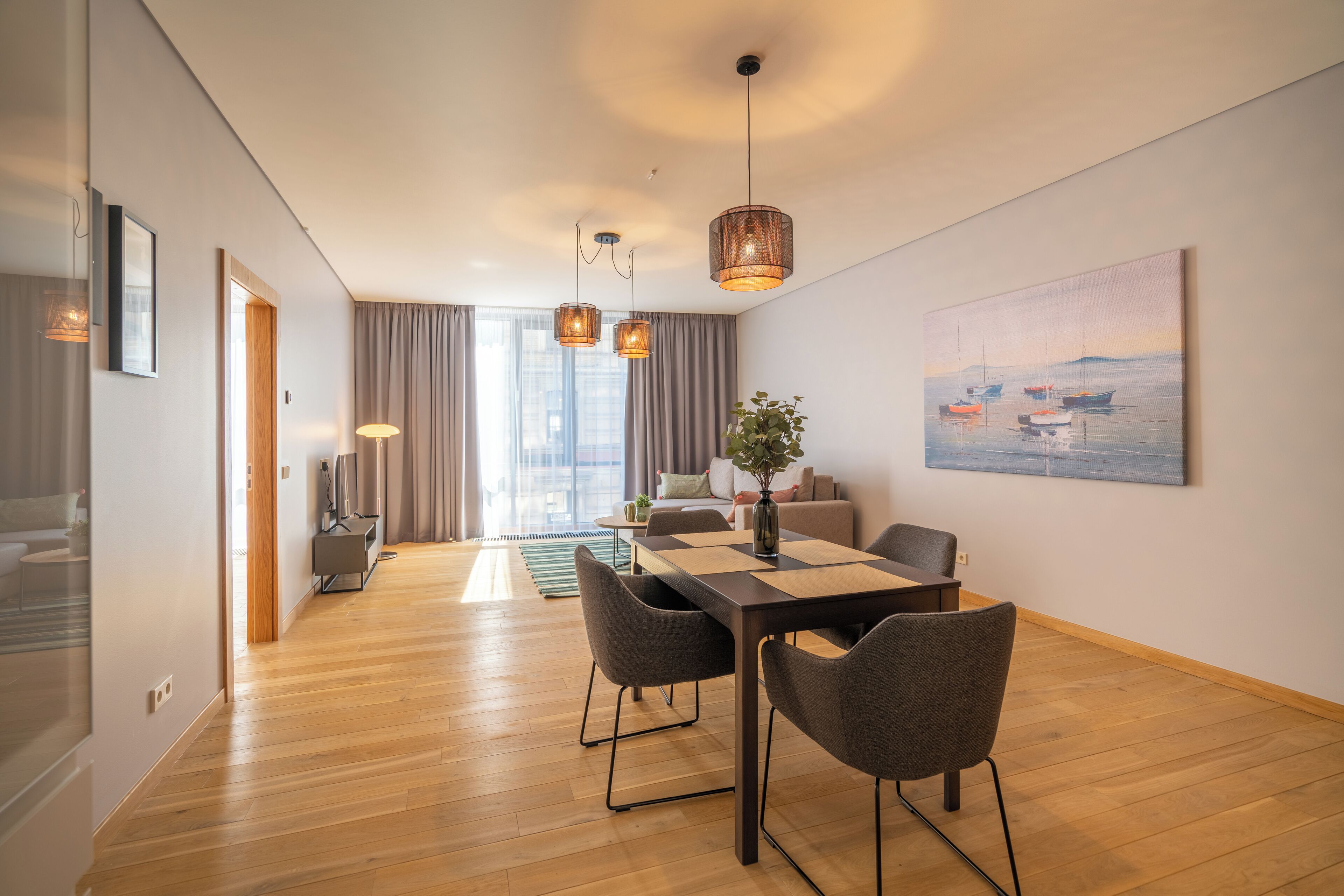 Una habitación de hotel moderna con piso de madera, un área de comedor con mesa y sillas, un sofá, un televisor y un cuadro con barcos en la pared.