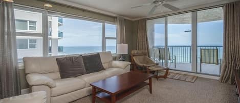 Un salón amplio con un sofá color beige, una mesa de centro de madera y una silla. Ventanas grandes ofrecen vistas al océano y un balcón.
