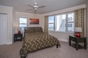 A bedroom with a bed, a ceiling fan, a window with curtains, and a painting on the wall. - Dimucci Twin Towers 1105 (Daytona Beach Shores)