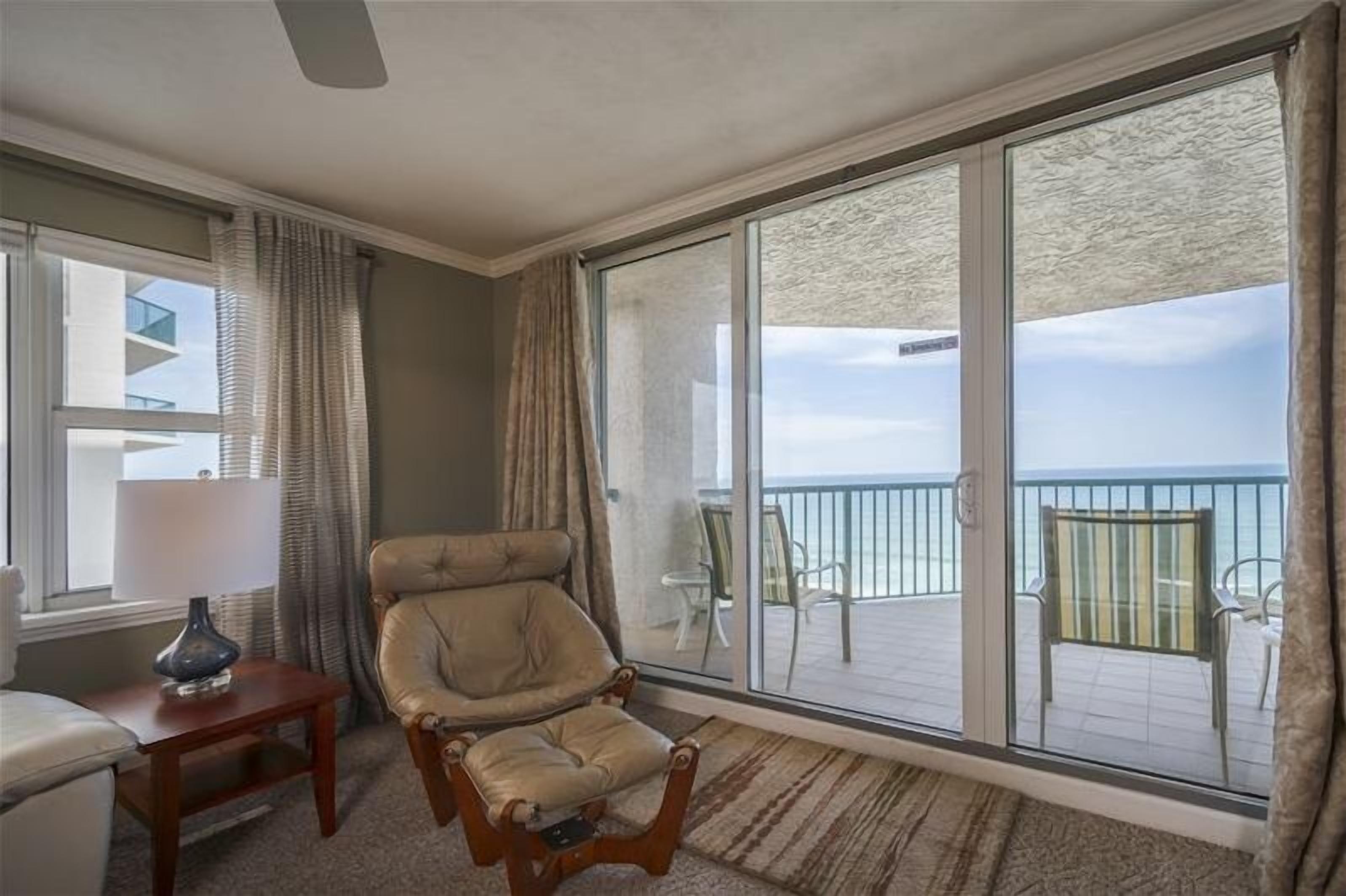 Habitación con un gran ventanal corredero que da a un balcón con vistas al océano. Hay una silla acolchada y una mesita con una lámpara.