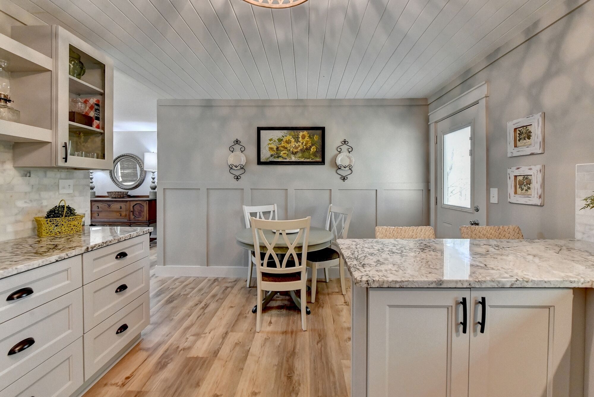 A kitchen with a wooden floor, white cabinets, a marble countertop, and a dining area with a table and chairs.