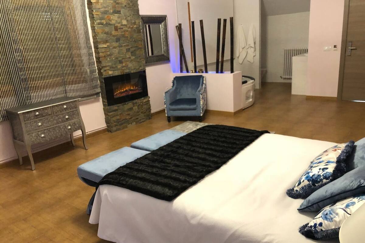 A bedroom with a bed, a stone fireplace, a blue armchair, and a white chest of drawers.
