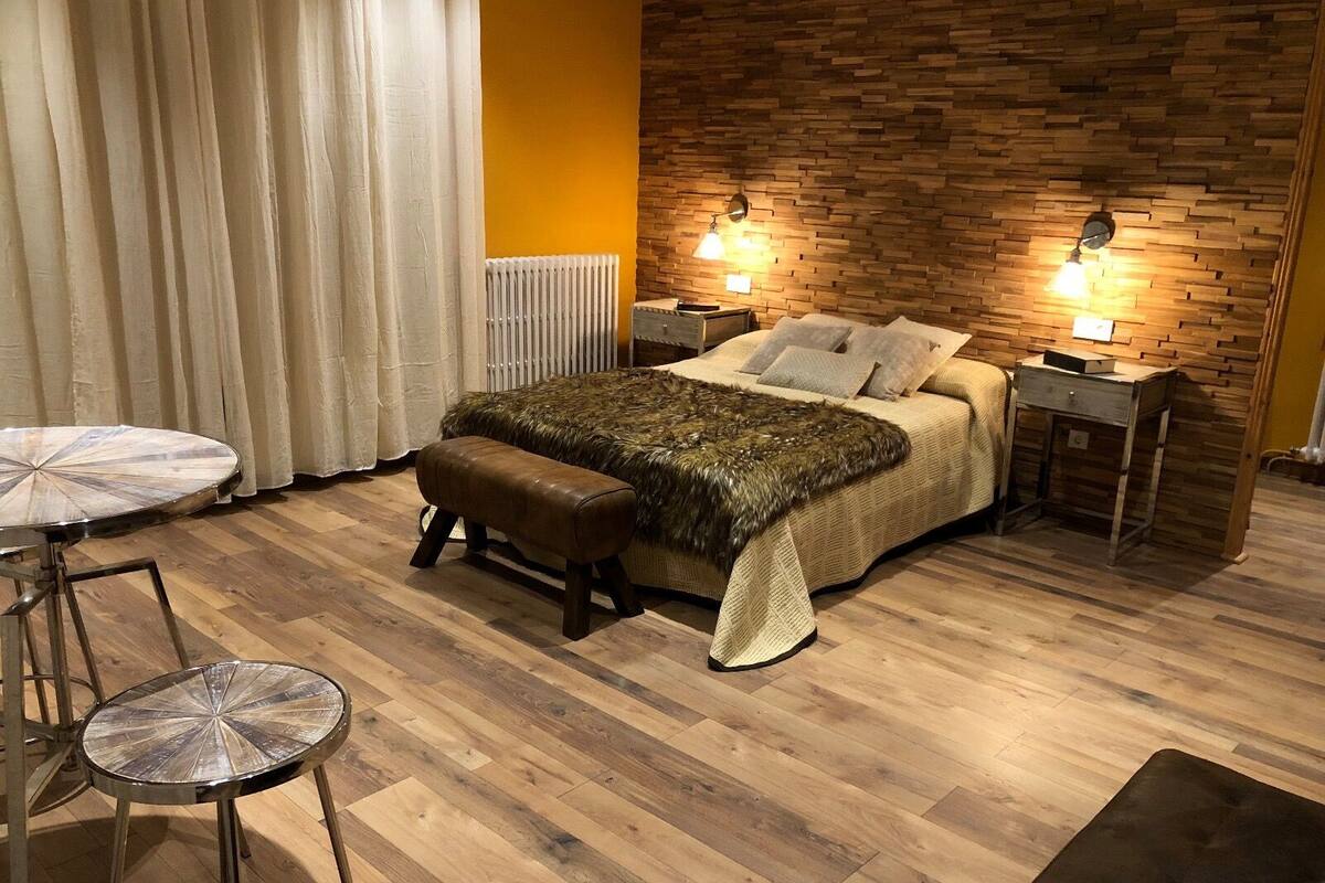 A bedroom with a wooden headboard, a bed with a fur throw, two bedside lamps, a wooden nightstand, a round table with a chair, and a window with curtains.