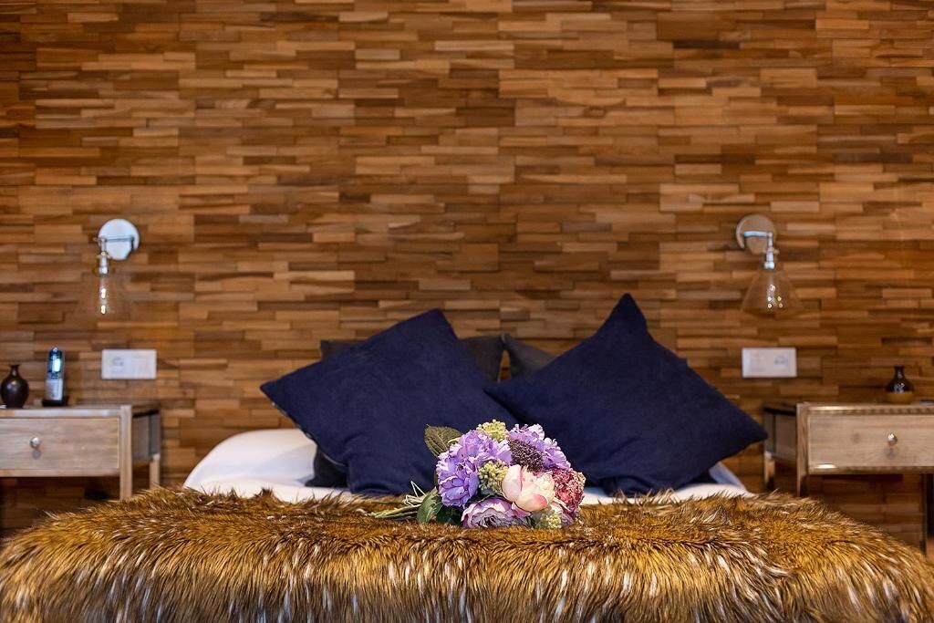 Un dormitorio con pared de madera, una cama con almohadas oscuras, una alfombra mullida y una mesita de noche con una lámpara.