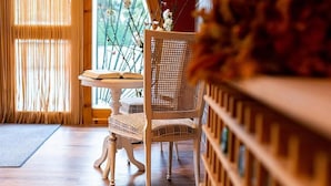 A room with a wooden floor, a wicker chair, a small table with a book, and a window with a view of greenery.