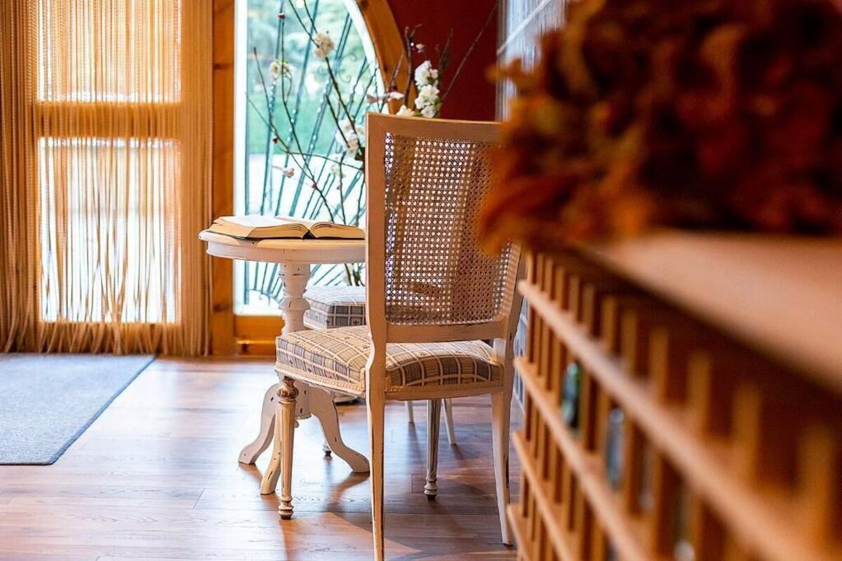 A room with a wooden floor, a wicker chair, a small table with a book, and a window with a view of greenery.