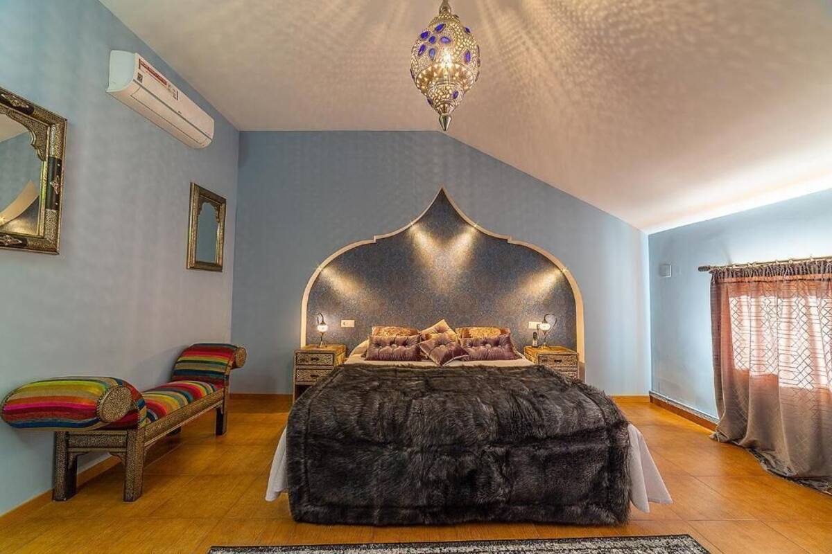 A bedroom with a bed, a bench, a chandelier, and a window with curtains.