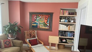 Ein Wohnzimmer mit einem beigefarbenen Sofa, einem Holzstuhl mit Kissen, einem Bücherregal voller Bücher, einer Topfpflanze und einem farbenfrohen abstrakten Gemälde an der Wand.