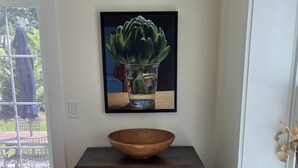 Ein Holzschrank mit einer Schüssel darauf, ein gerahmtes Bild einer Pflanze in einem Glas Wasser und ein Fenster mit Blick auf Gartenmöbel.