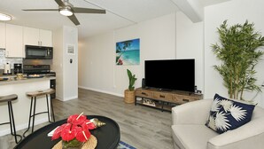 A modern living room with a flat-screen TV, a ceiling fan, a dining area with stools, and a kitchen with a microwave.