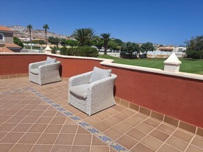 Eine Terrasse mit zwei Korbstühlen, Fliesenboden und Blick auf eine grüne Landschaft mit Bäumen und Gebäuden in der Ferne.