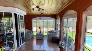 Amplia zona de patio con mobiliario de mimbre, un ventanal de vidrio que da al exterior y vista a una piscina.