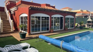 Una casa con piscina, un patio con sillas blancas y un césped verde.