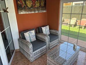 A wicker sofa with cushions, a glass-top table, and a view of a green lawn through a glass door.