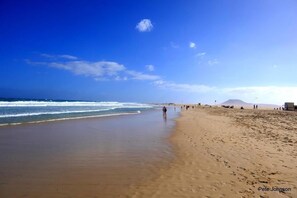 A wide sandy beach with gentle waves, a clear sky, and a distant mountain.