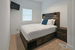 A bedroom with a bed, a nightstand, a wooden headboard, a TV mounted on the wall, and a window with blinds.