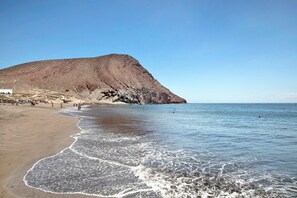 Una playa con una gran colina al fondo, cielos despejados y agua tranquila.