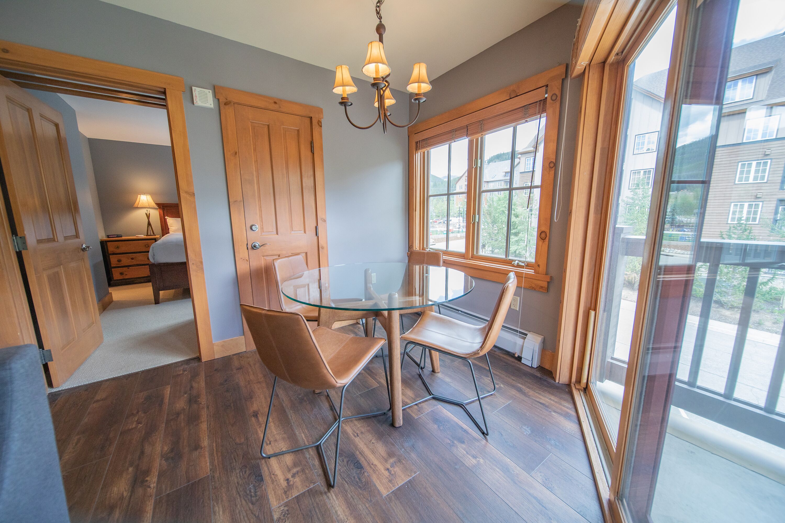 A room with a glass table, two chairs, a chandelier, wooden flooring, and large windows.
