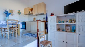 A kitchen with wooden cabinets, a dining table with chairs, a white cabinet with shelves, and a blue vase on the wall.