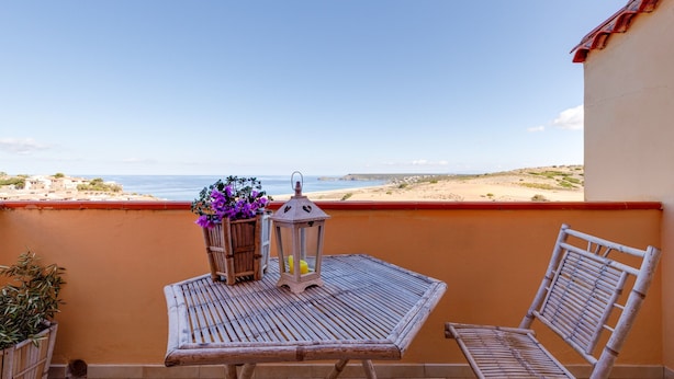 A balcony with a table, two chairs, a lantern, and a flower vase overlooking a beach.