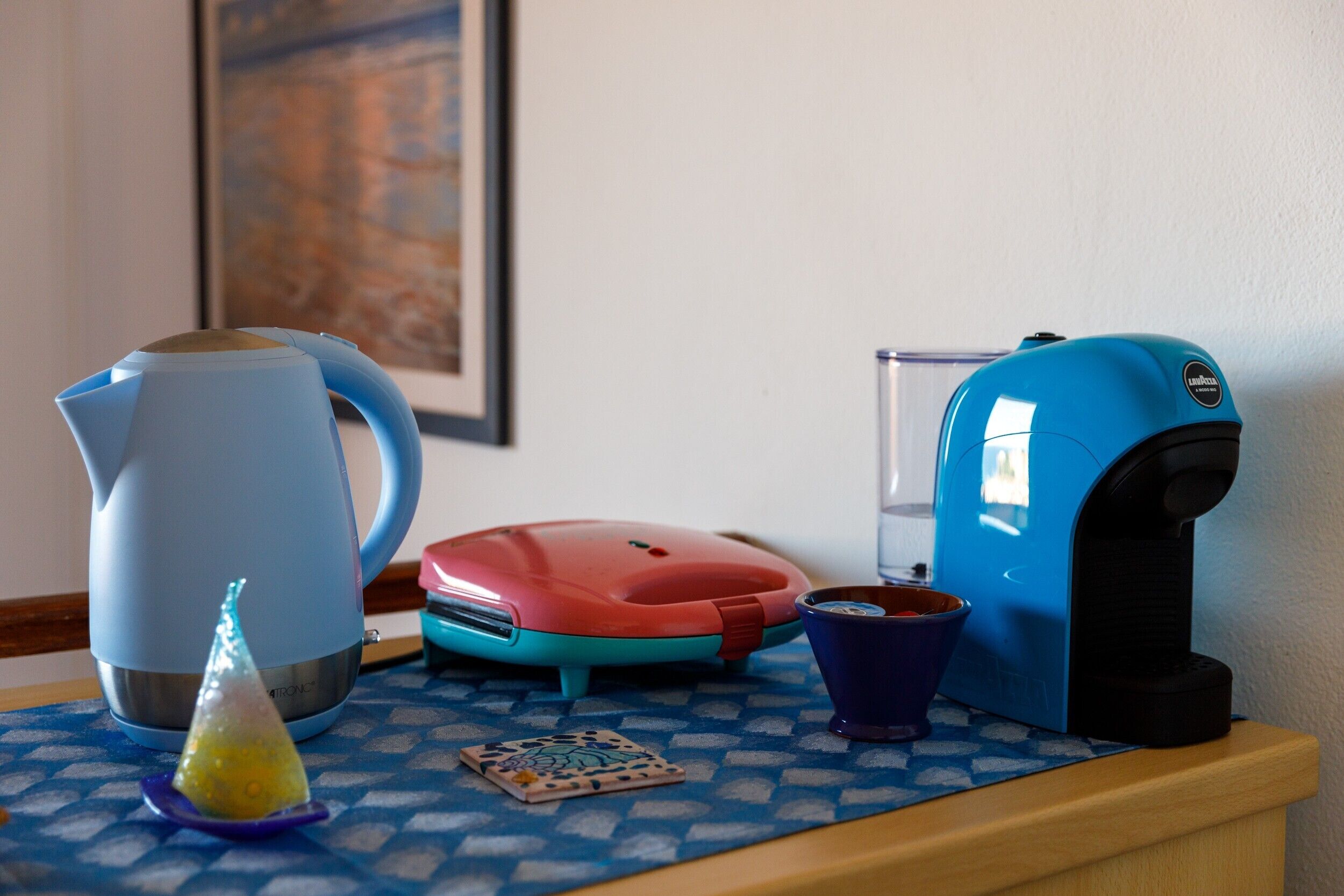 Un plan de travail de cuisine avec une bouilloire, une machine à café, un appareil de cuisine rouge et une tasse bleue.