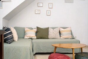 Een woonkamer met een hoekbank, een ronde houten salontafel en een rode poef. Er hangen vier ingelijste foto’s aan de muur.