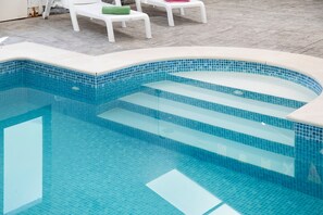Ein Schwimmbad mit klarem, blauem Wasser, mit Fliesenstufen und weißen Liegestühlen mit grünen Kissen.