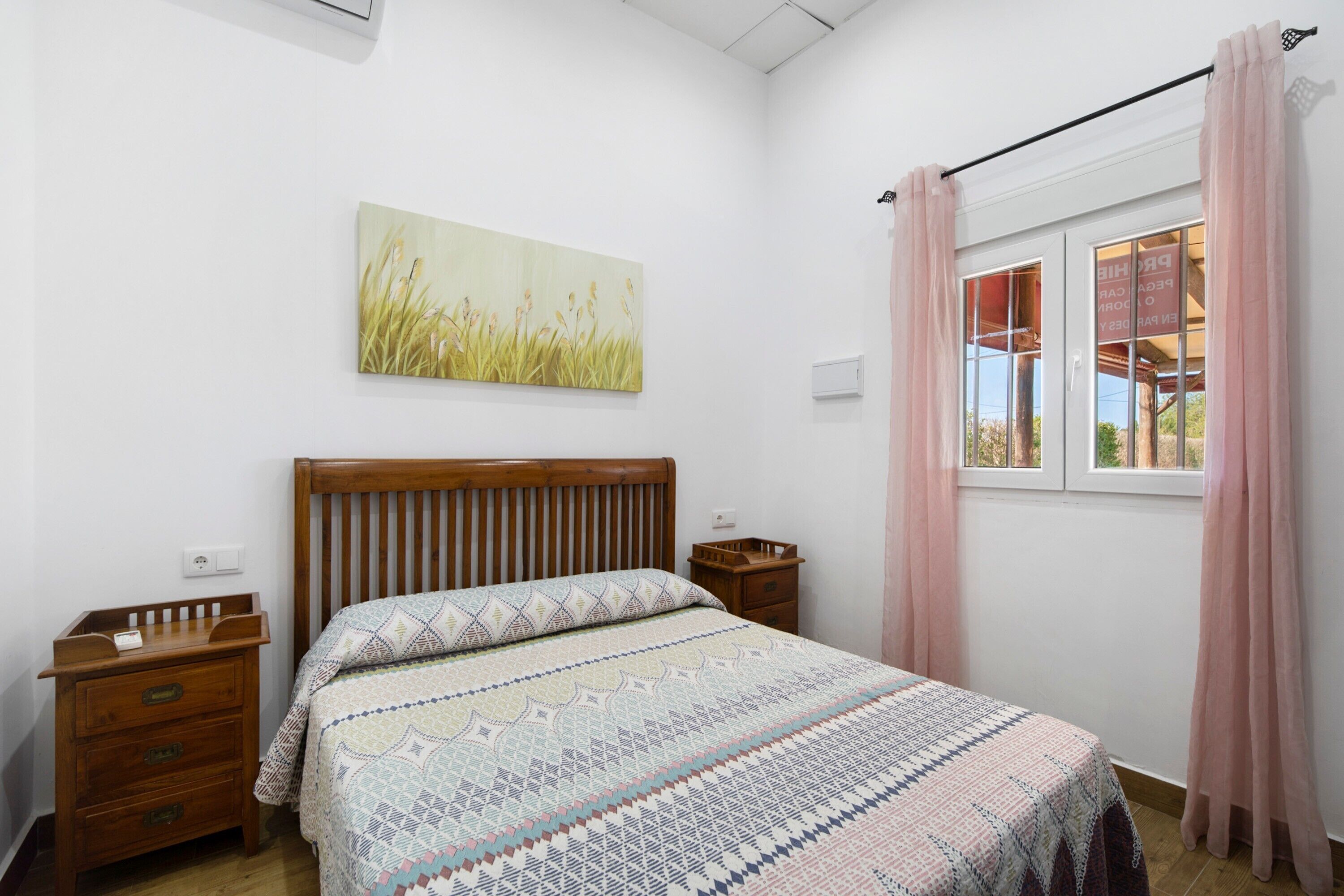 Un dormitorio con cama de madera, mesita de noche, ventana con cortinas y un cuadro colgado en la pared.