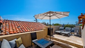 A rooftop patio with terracotta tiles, a table with a glass top, two lounge chairs, a large umbrella, and a decorative lantern.