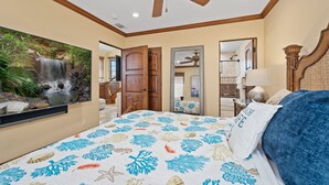 A bedroom with a bed, a ceiling fan, a large wall-mounted TV, a mirror, and a door leading to another room.