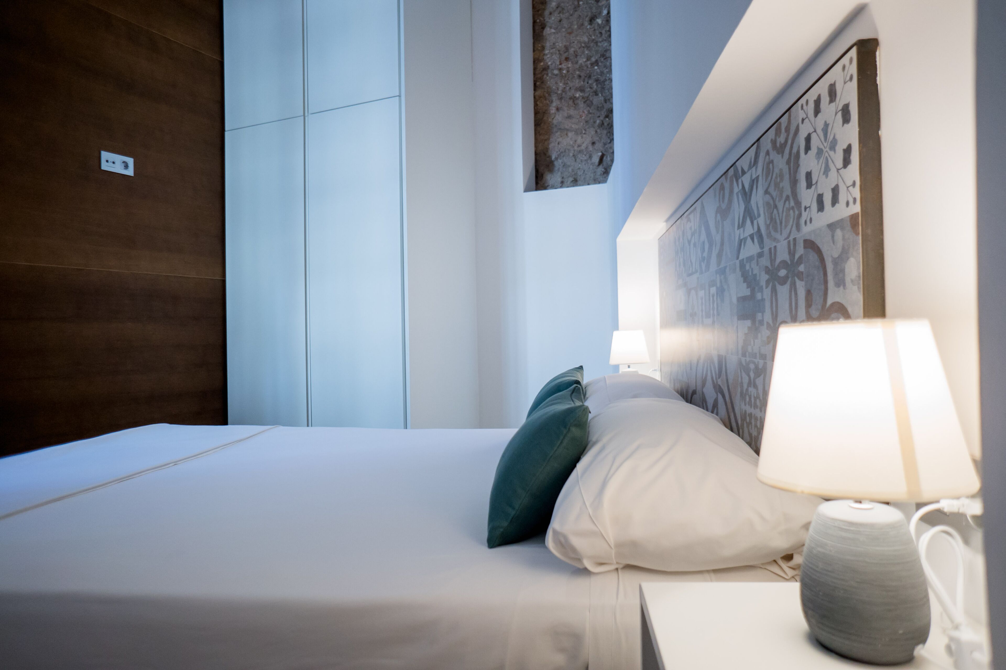 A neatly made bed with white bedding and green and white pillows, a bedside lamp, and a wall with a patterned design.