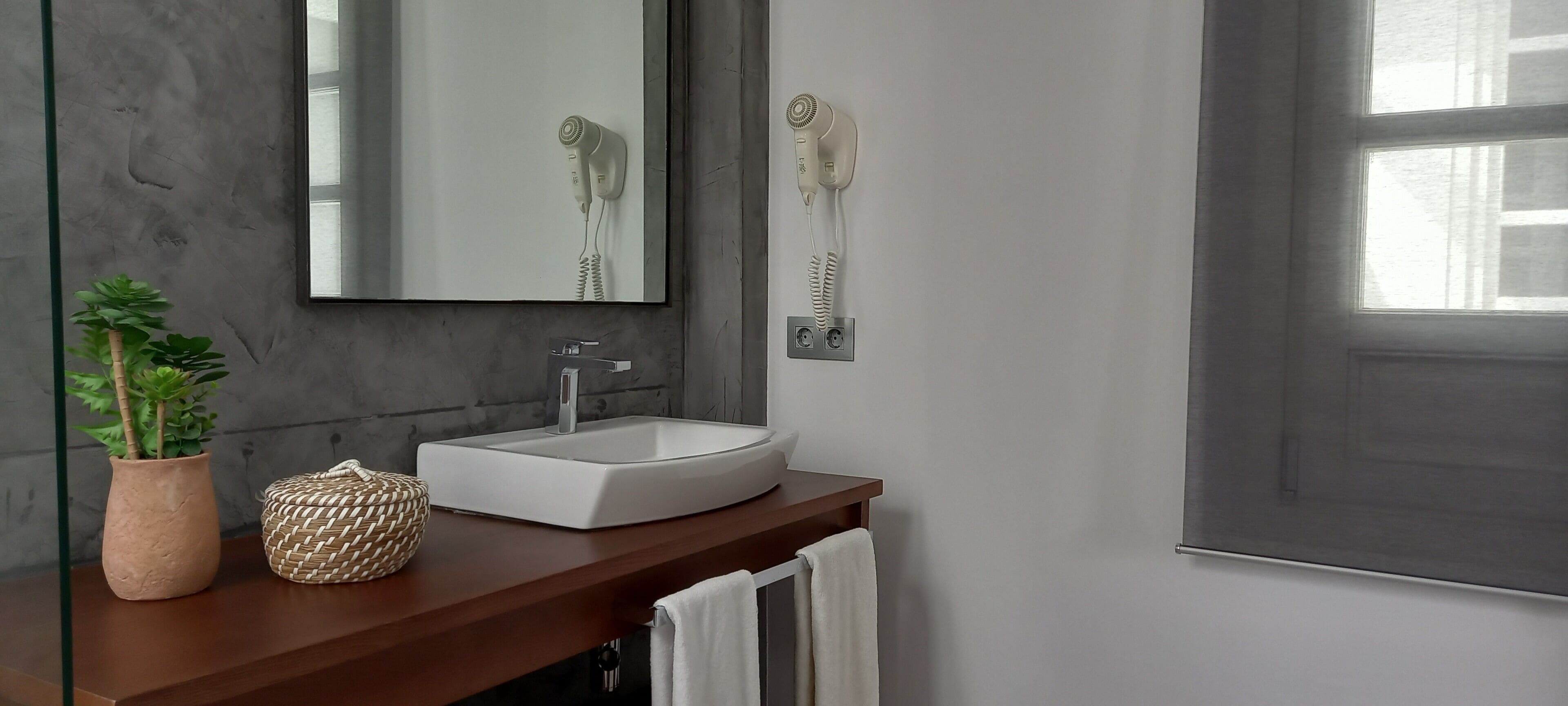 A bathroom with a white sink, a mirror, a hairdryer, a potted plant, and a woven basket on the counter.