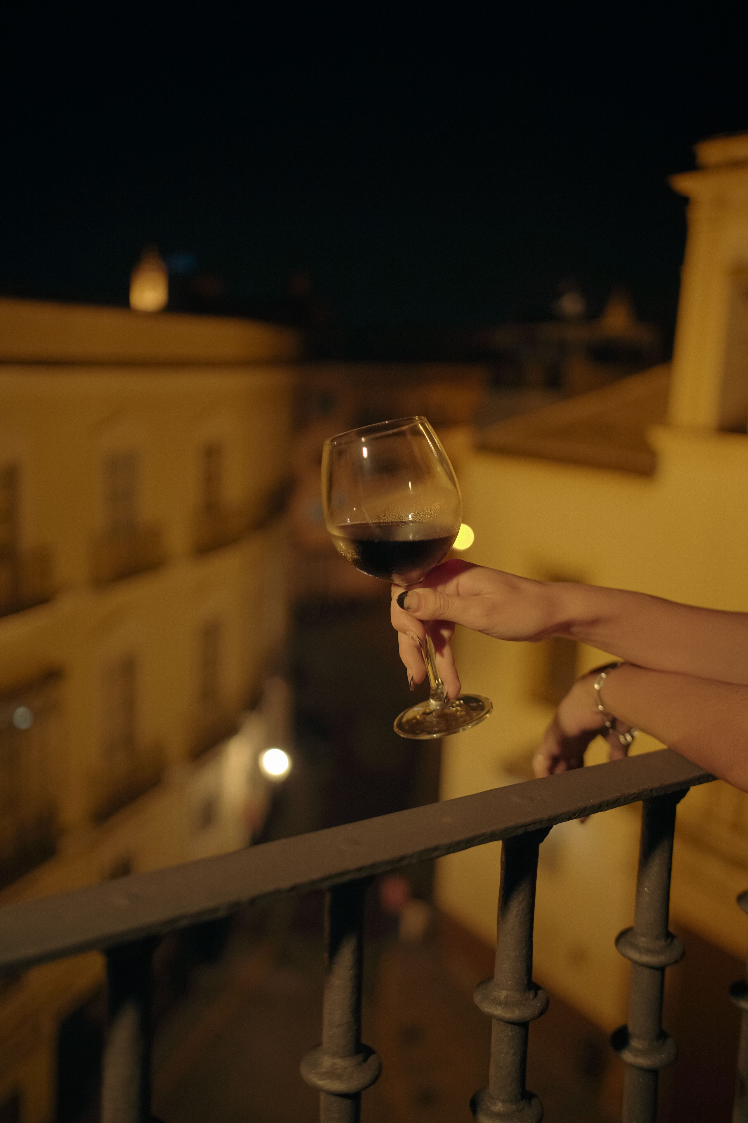 Une main tenant un verre à vin, avec une rambarde et des bâtiments en arrière-plan.