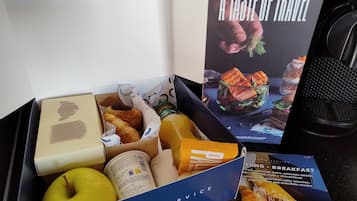 Une boîte pour le petit-déjeuner contenant divers aliments, notamment des viennoiseries, une bouteille de jus de fruits et un pot de confitures ou de pâte à tartiner.