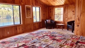 Una habitación de cabaña de madera con una cama grande, dos ventanas, una silla y una pequeña mesa.